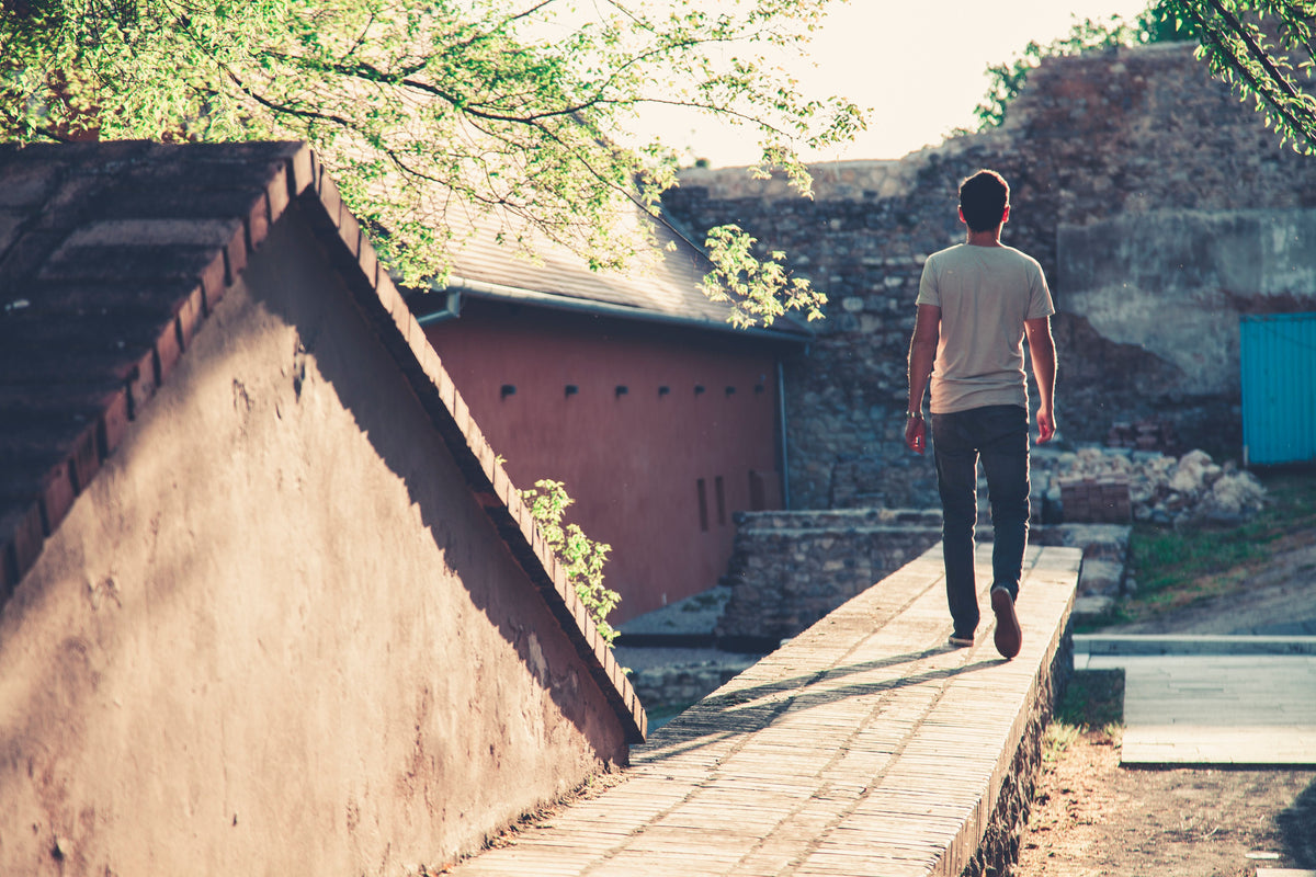 Man in a gray t-shirt and black denim jeans walking along a sunlit urban pathway, representing the timeless, everyday wearability of quality pre-owned men's vintage jeans