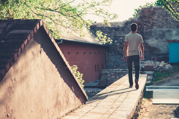 Man in a gray t-shirt and black denim jeans walking along a sunlit urban pathway, representing the timeless, everyday wearability of quality pre-owned men's vintage jeans
