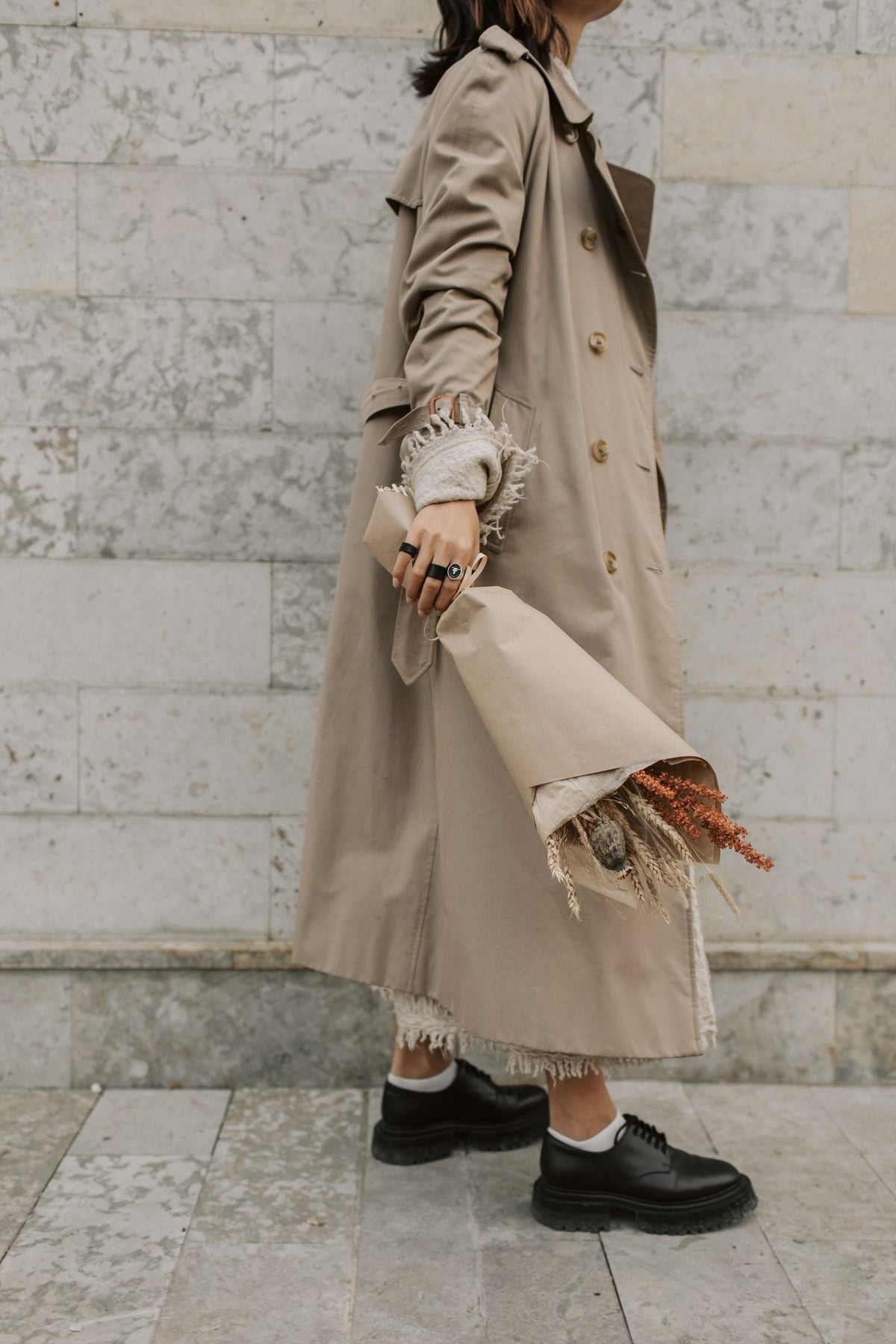 Camel trench coat styled with dried florals against a marble wall in soft autumn light