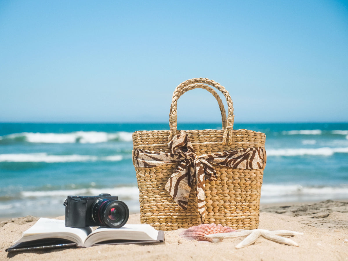 Woven beach bag, book, and seashells arranged on sand near the ocean