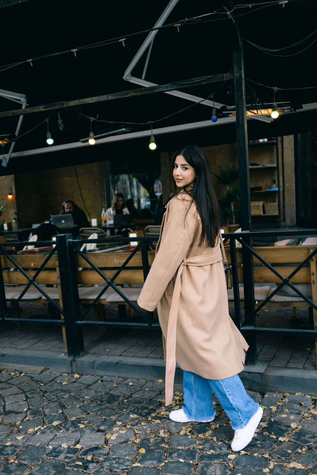 Woman in a beige trench coat walking on a cobblestone pavement, representing the timeless silhouette at the center of CENT'ANNII's pre-owned trench coat buying guide
